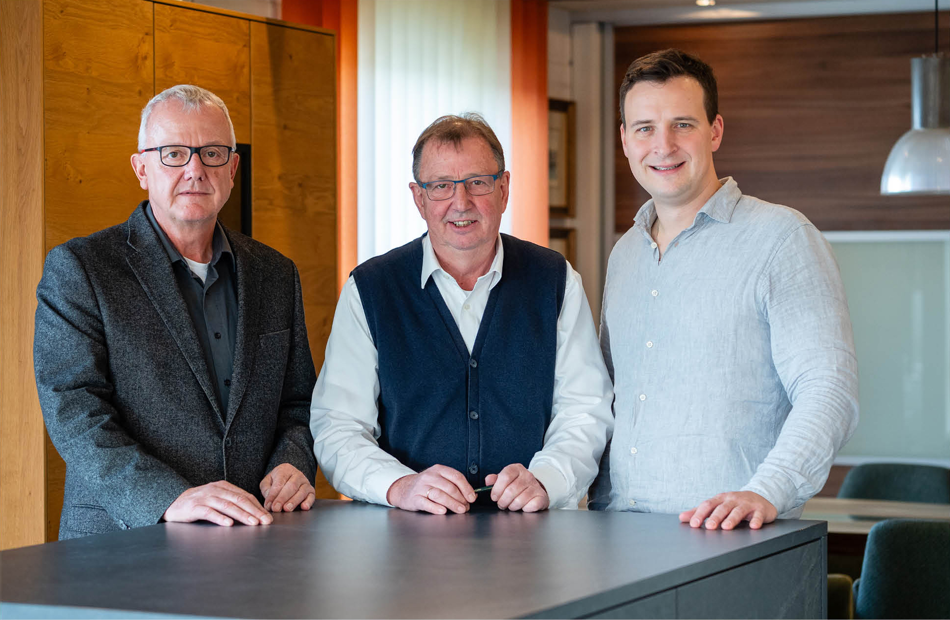 Die drei Geschäftsführer stehen vor einem Tisch und lächeln in die Kamera.